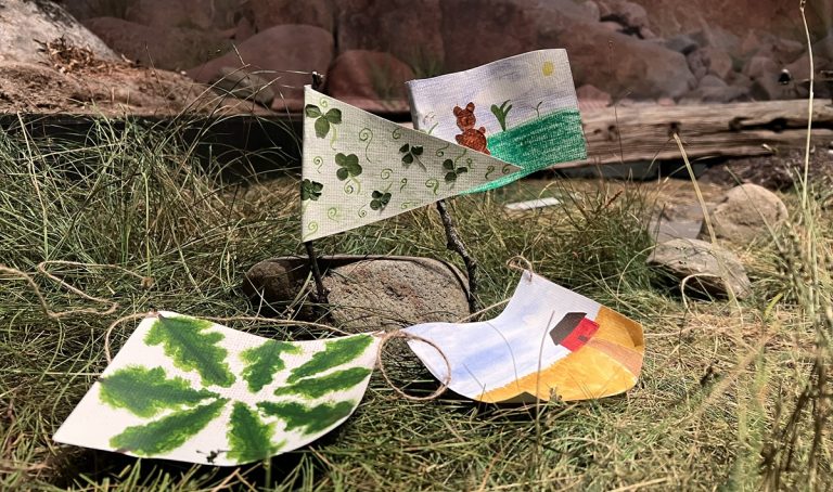 Neljä erilaista, värikkäästi koristeltua, paperista askarreltua lippua sijoiteltuna Arkin näyttelyyn ruohon ja kivien päälle.